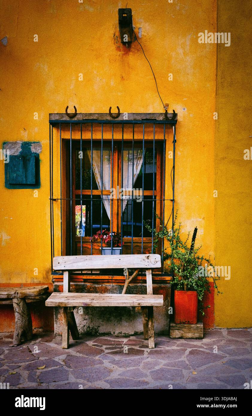 Una scena affascinante caratterizzata da una panchina rustica in legno di fronte ad una parete gialla e da una finestra adornata con piante e bar Foto Stock