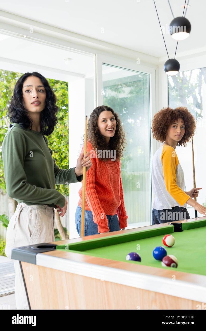 Tre amiche in piedi intorno al tavolo da biliardo in una luminosa sala giochi che regge segnali e palle da biliardo Foto Stock