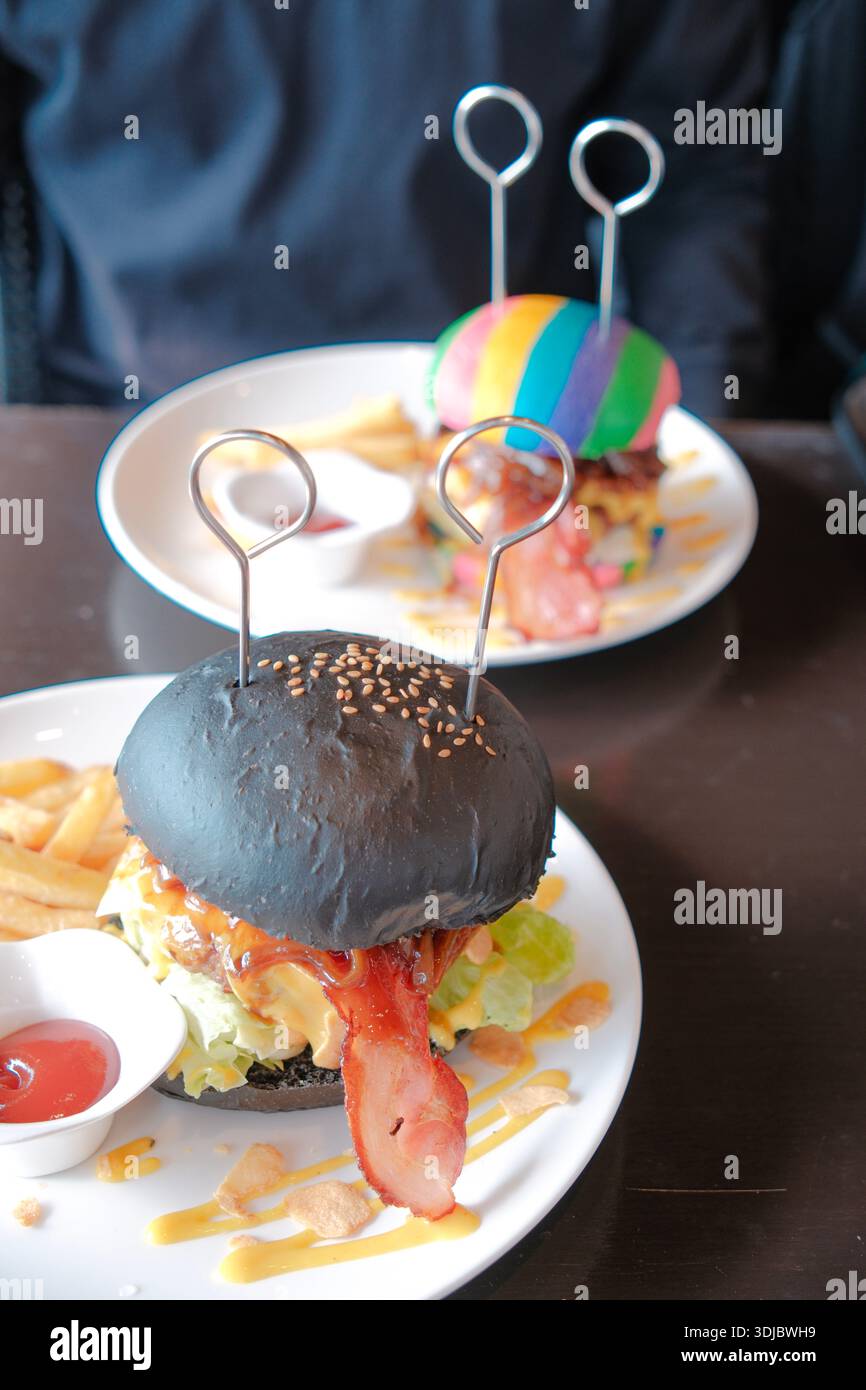 Hamburger gourmet fatto a mano con panino nero, pancetta, formaggio e verdure fresche serviti con patatine fritte su un piatto. Foto Stock
