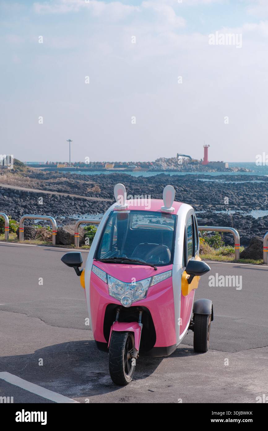 Uno scooter elettrico rosa a tre ruote è parcheggiato lungo una strada costiera, con la costa rocciosa e il mare visibili sullo sfondo. Foto Stock