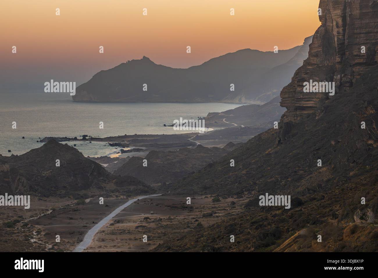 Un'ampia vista aerea della spettacolare costa dell'Oman al tramonto. Una strada tortuosa serpeggia lungo la riva, fiancheggiata da aspre montagne e dalla tranquilla A Foto Stock