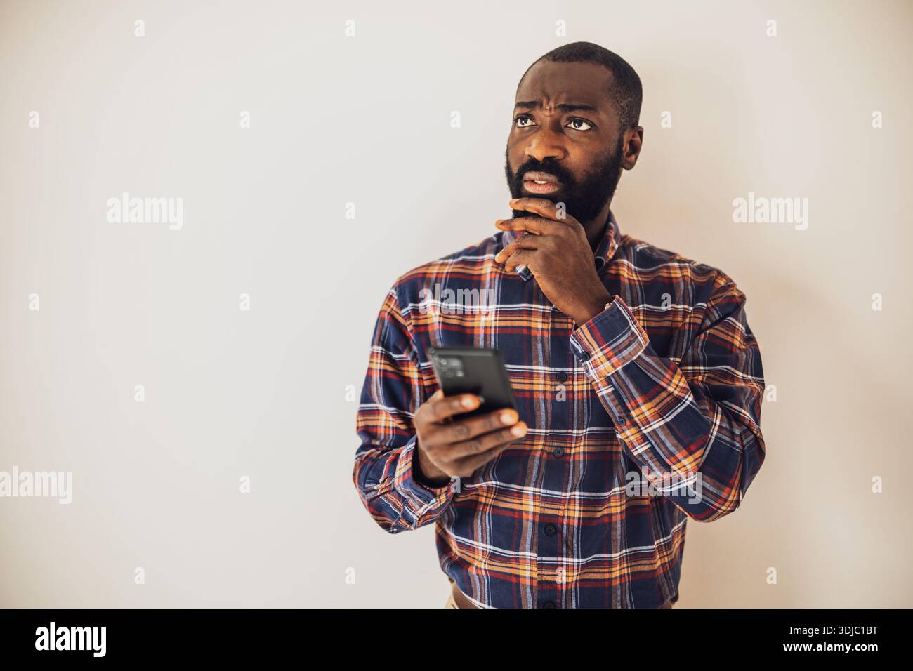 Uomo nero ansioso in una camicia accogliente davanti alla parete e scrivendo messaggi sullo smartphone. Foto Stock