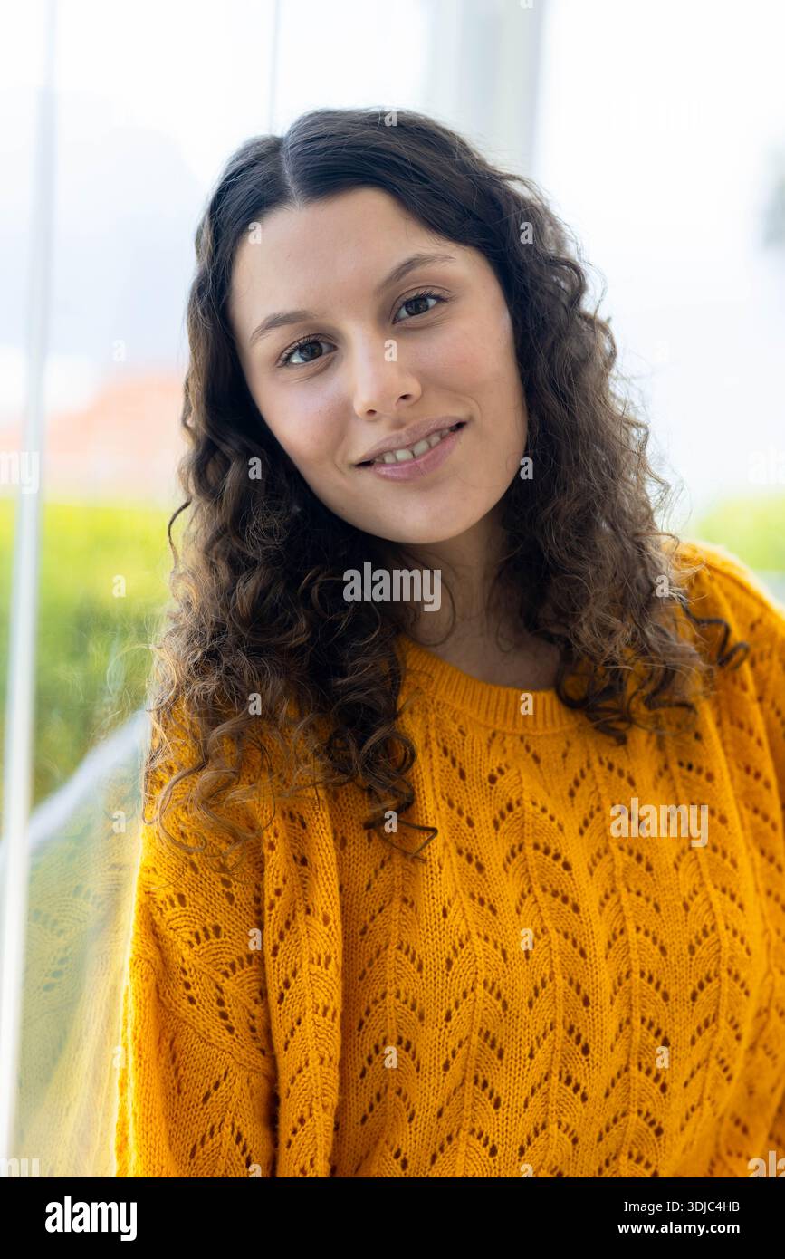 Donna di 20 anni, appoggiata vicino a una grande finestra di vetro, indossa un maglione in maglia giallo senape Foto Stock