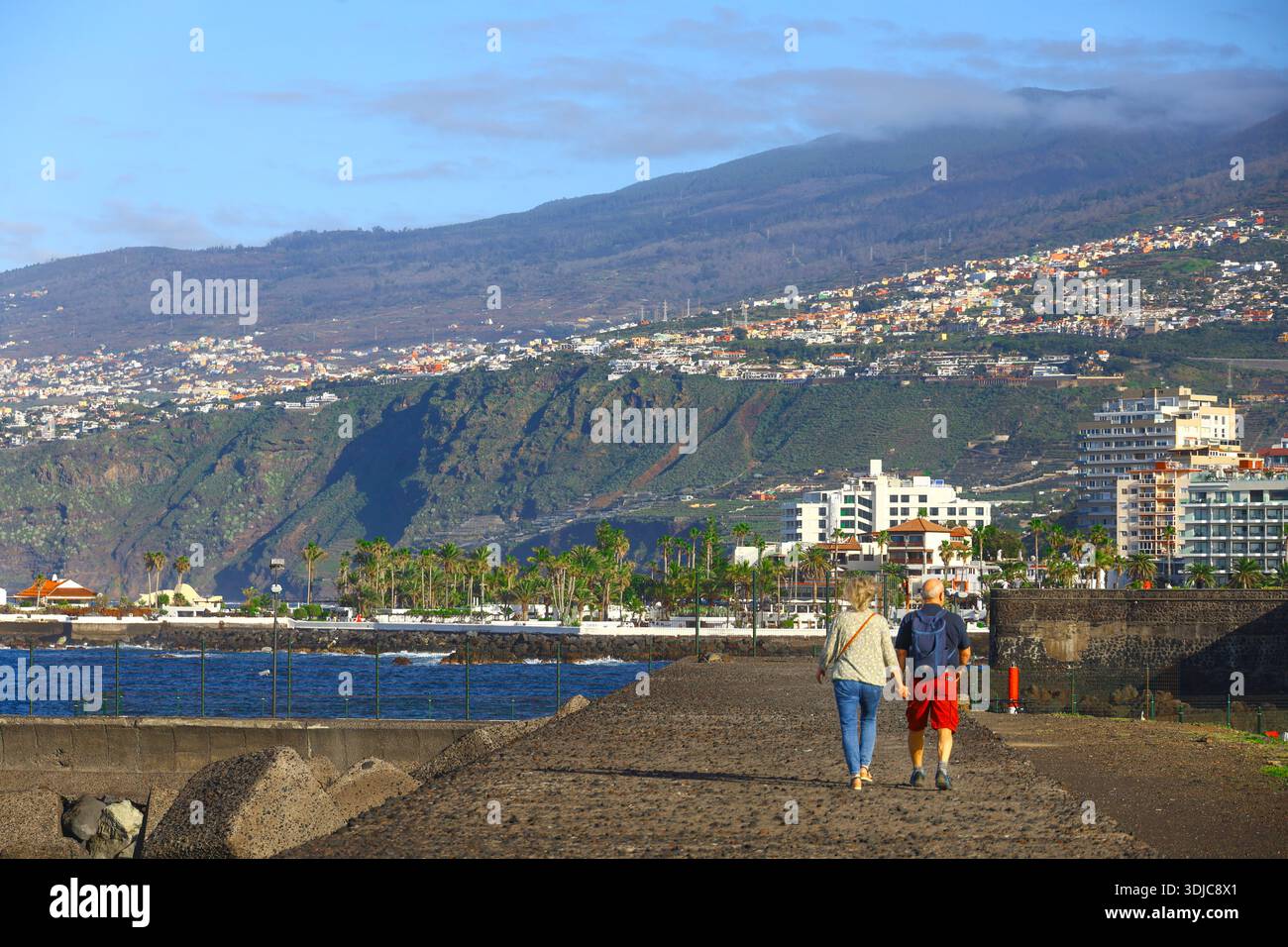 Puerto de la Cruz, 01152026 - coppia che cammina lungo la passeggiata costiera con paesaggio urbano e vista sull'oceano Foto Stock