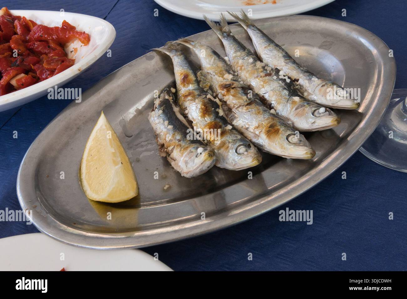 Primo piano di sardine appena grigliate condite con sale, servite su un piatto di metallo ovale rustico con un spicchio di limone giallo brillante. Sullo sfondo, a Foto Stock