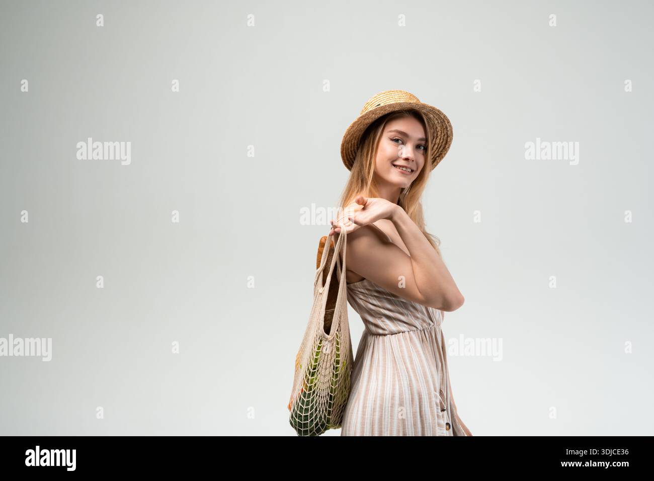 Una giovane donna sorridente che indossa un cappello di paglia e un leggero abito estivo tiene una borsa della spesa riutilizzabile su uno sfondo neutro. L'immagine rappresenta un negozio sostenibile Foto Stock