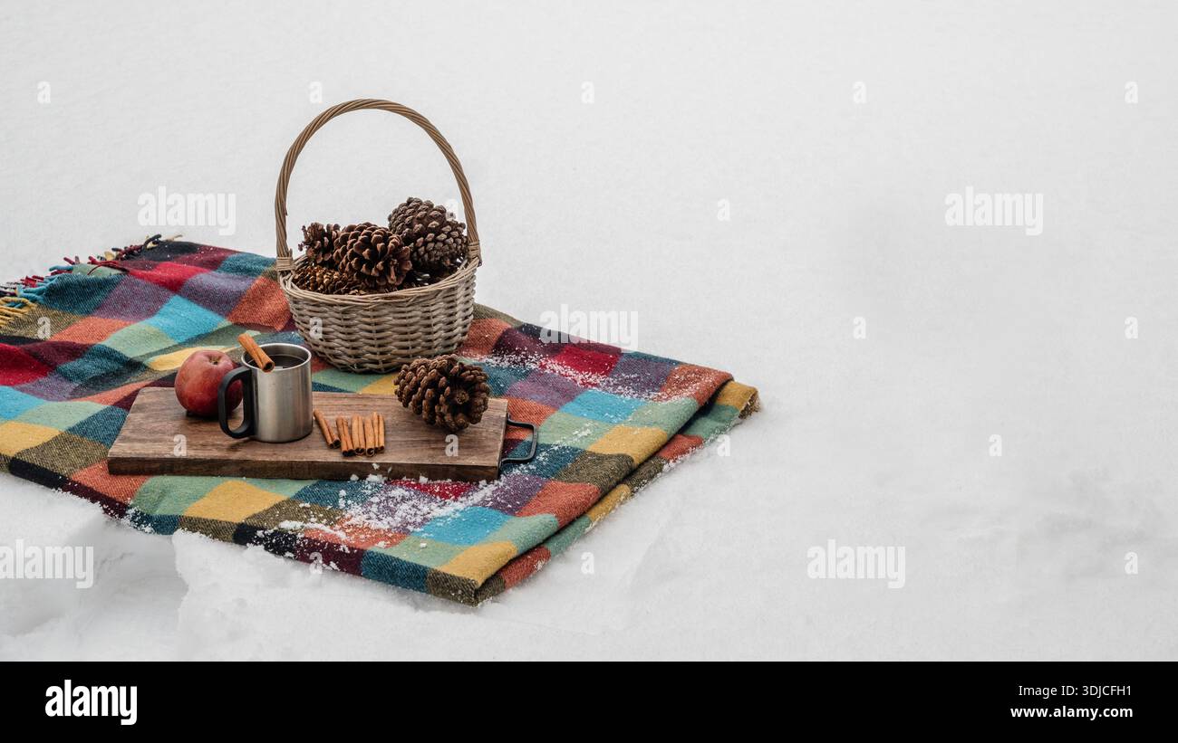 Picnic invernale con coperta, cestino, coni di pino e bevanda calda sulla neve. Accogliente stile di vita all'aperto e concetto stagionale. Foto Stock