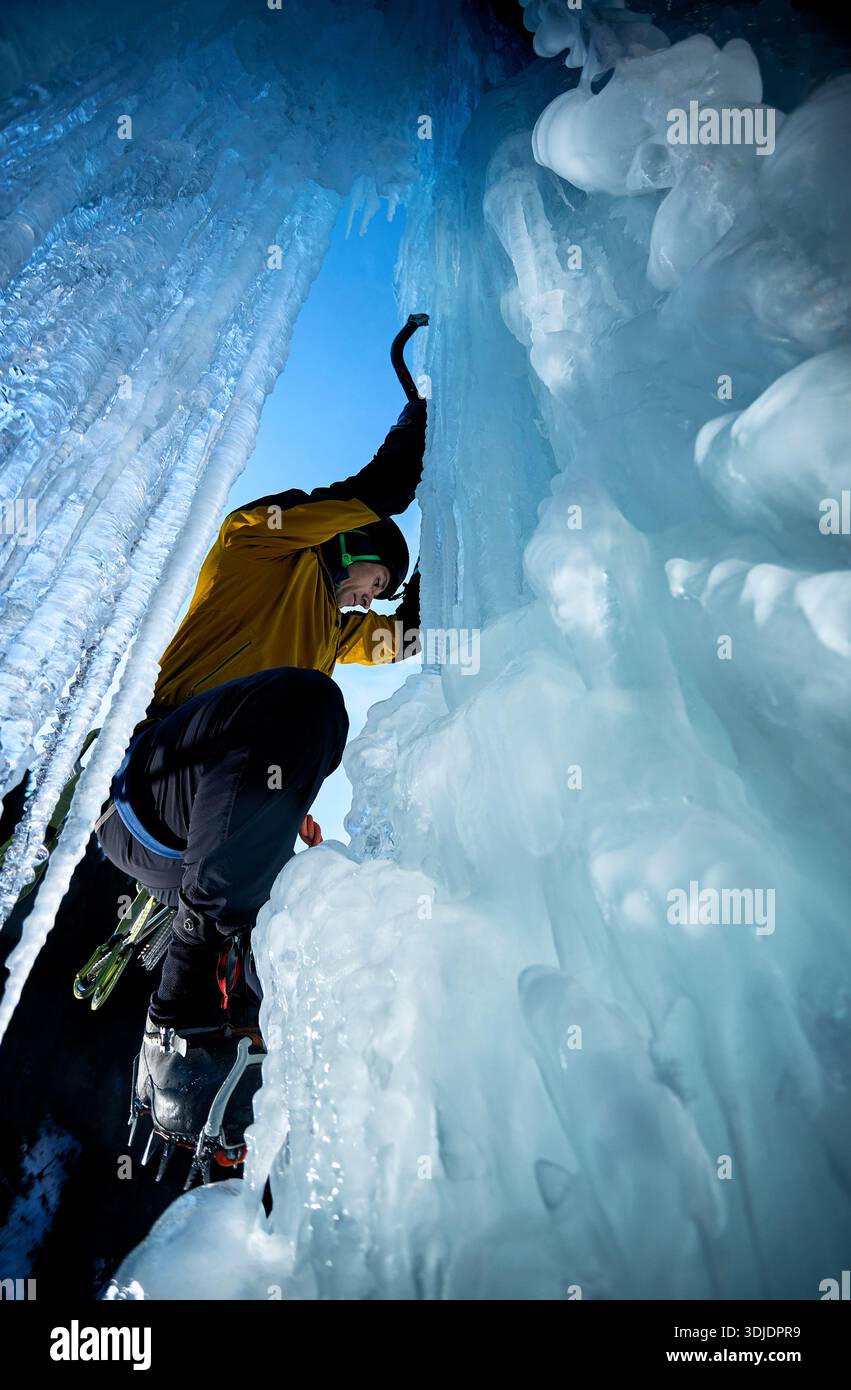 L'uomo con la giacca gialla si arrampica su una parete ghiacciata alla grande cascata con un'ascia e ramponi contro lo sci blu nelle montagne invernali. Sport alpinista Foto Stock