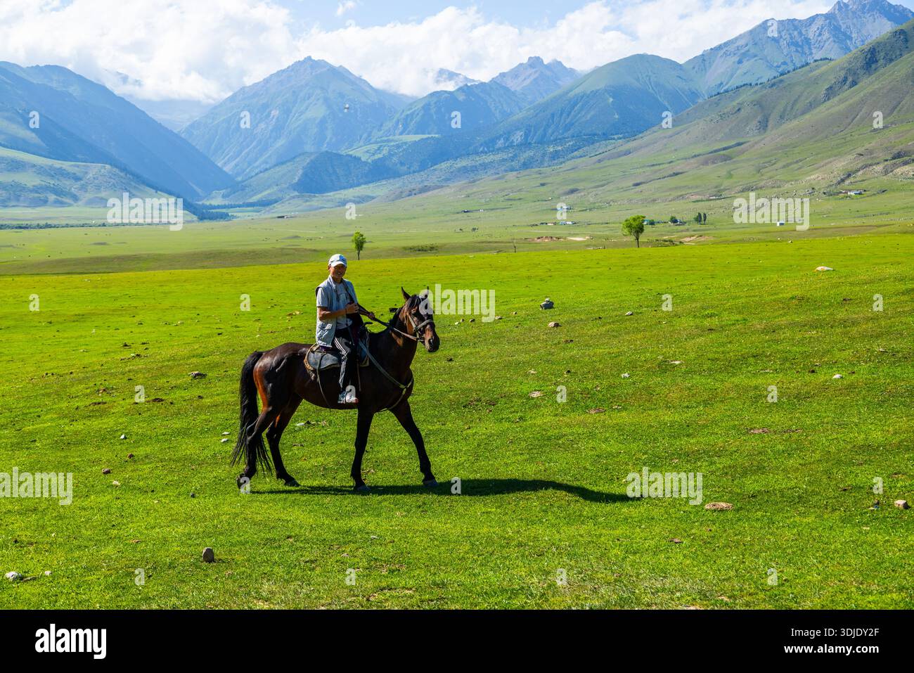 Un uomo adulto sorride mentre cavalca un cavallo bruno in una pianura erbosa. Lo sfondo panoramico delle montagne aggiunge un'atmosfera serena e avventurosa. Semenovskoye Gorge, Kirghizistan - 17 luglio 2024 Foto Stock