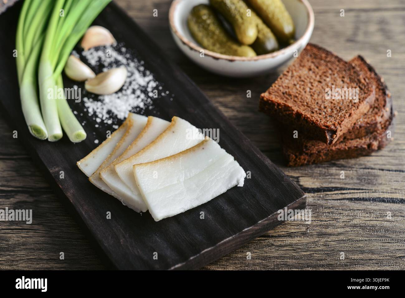 Maiale fatback su un tagliere di legno con fette di pane di segale, aglio, cetrioli sottaceto in un piatto e cipolle verdi fresche, primo piano Foto Stock