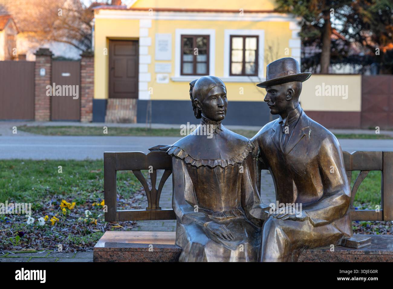 VINKOVCI, CROAZIA, 12.27.2025: Statua di bronzo di un uomo e di una donna seduti su una panchina del parco, adagiata su uno sfondo giallo brillante. Esprime romanticismo, Foto Stock