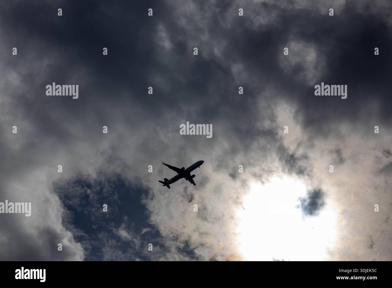 Una potente silhouette a basso angolo di un jet passeggeri che attraversa un paesaggio nuvoloso, scuro e turbolento. Il sole luminoso è parzialmente oscurato da t Foto Stock