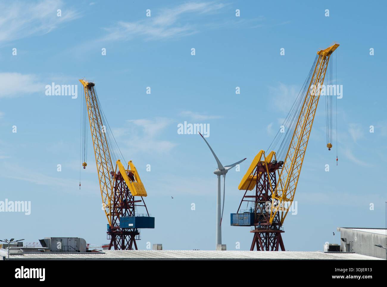 Gru gialle industriali in piedi nel porto marittimo sotto il cielo blu con turbina eolica. Grandi attrezzature portuali per il trasporto di merci per attività di spedizione e logistica. Marit Foto Stock