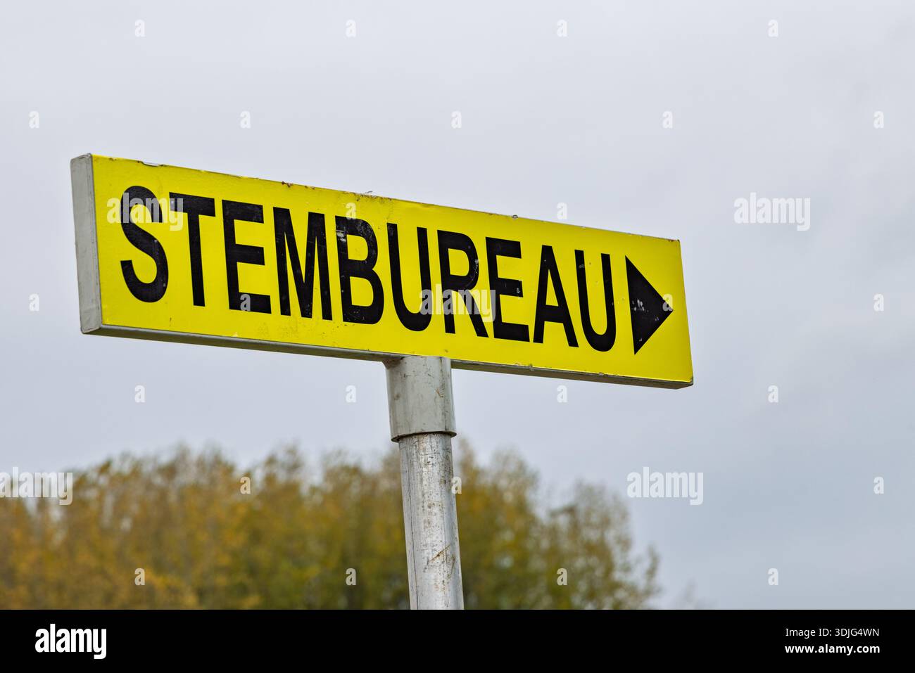 Un cartello giallo olandese per una stazione elettorale (Stembureau) con una freccia nera, posto contro un cielo nuvoloso e alberi autunnali offuscati. Foto Stock
