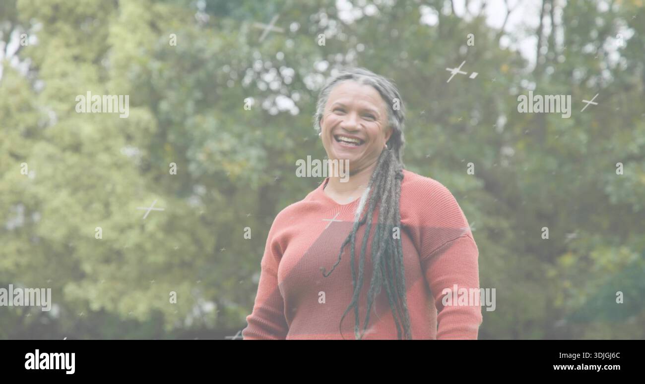 Donna anziana sorridente che indossa un maglione ruggine e mostra lunghi abiti nel parco, con piccole croci bianche Foto Stock