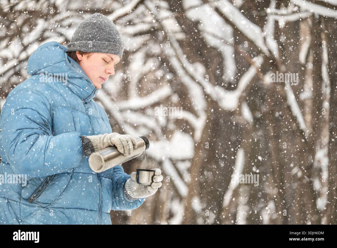 Giovane uomo in giacca invernale che versa una bevanda calda dalla tazza thermos all'aperto durante le nevicate. Bevande calde nelle giornate fredde, stile di vita invernale e attività all'aperto Foto Stock