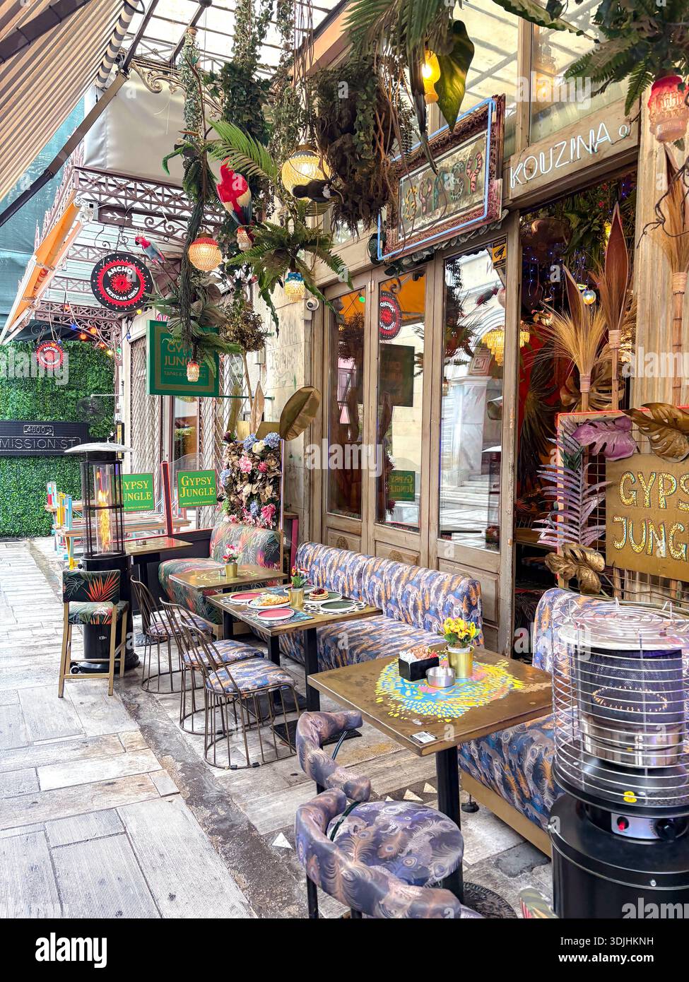 La giungla zingara, Kouzina. Atene, Grecia. Eclettica area pranzo all'aperto con vegetazione lussureggiante, vivaci posti a sedere a motivi geometrici e riscaldatori da patio. Foto Stock