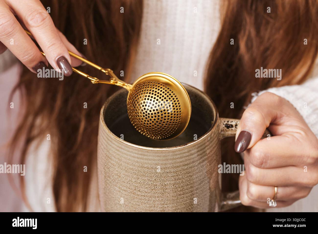 Donna che tiene in mano l'infusore di tè metallico sopra la tazza di ceramica durante la preparazione del tè a foglia sciolta. Primo piano delle mani che preparano una bevanda calda a casa, mostrando il rituale del tè. Foto Stock
