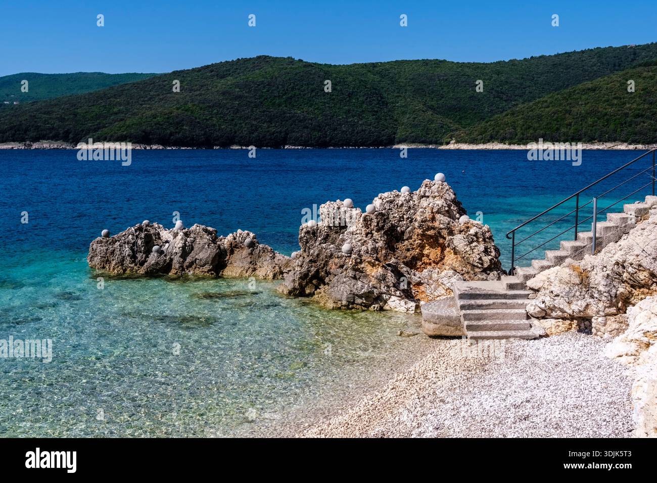 I gradini di pietra conducono a una spiaggia di ciottoli con formazioni rocciose nel mare turchese. Montagne boscose sono visibili in lontananza sotto un cielo limpido. Foto Stock
