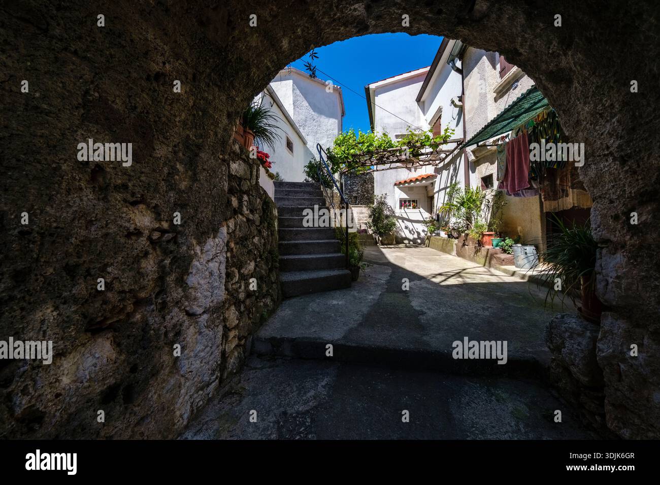 Ammira attraverso un arco in pietra uno stretto cortile con scale e case tradizionali. Piante verdi e un cielo blu sono visibili in lontananza. Foto Stock