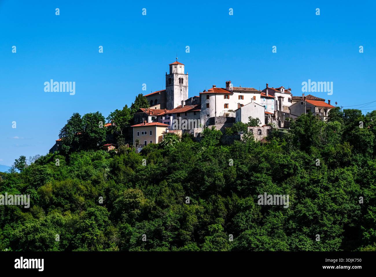 Vista del villaggio in cima alla collina di Mošćenice. Die Kirche St. Andrew si erge sopra le case circondato da alberi verdi sotto un cielo azzurro. Foto Stock