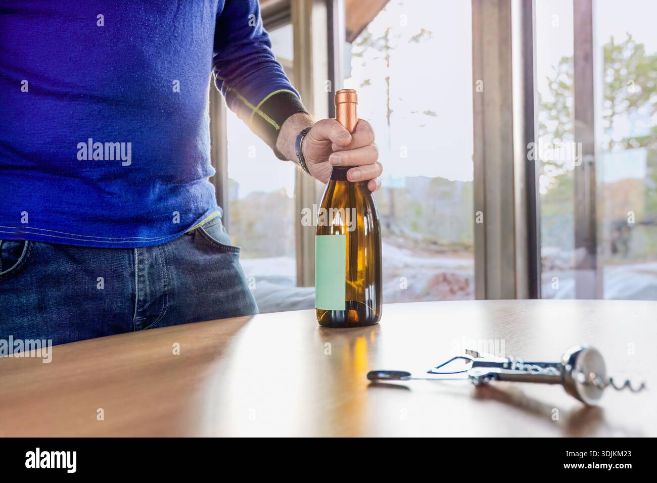 Uomo in maglione blu che stappa una bottiglia di vino su un tavolo di legno, con un cavatappi accanto, creando un'atmosfera accogliente per incontri sociali e celebrazioni Foto Stock