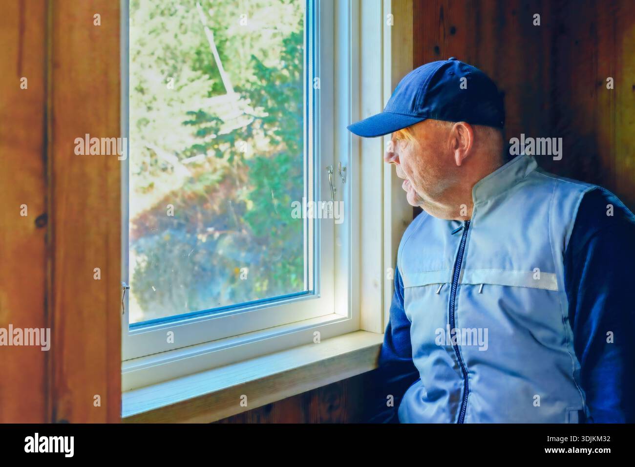Un uomo con cappello e giacca blu guarda fuori dalla finestra, circondato da pareti di legno, catturando un momento di riflessione e connessione con la natura e tranquillo Foto Stock