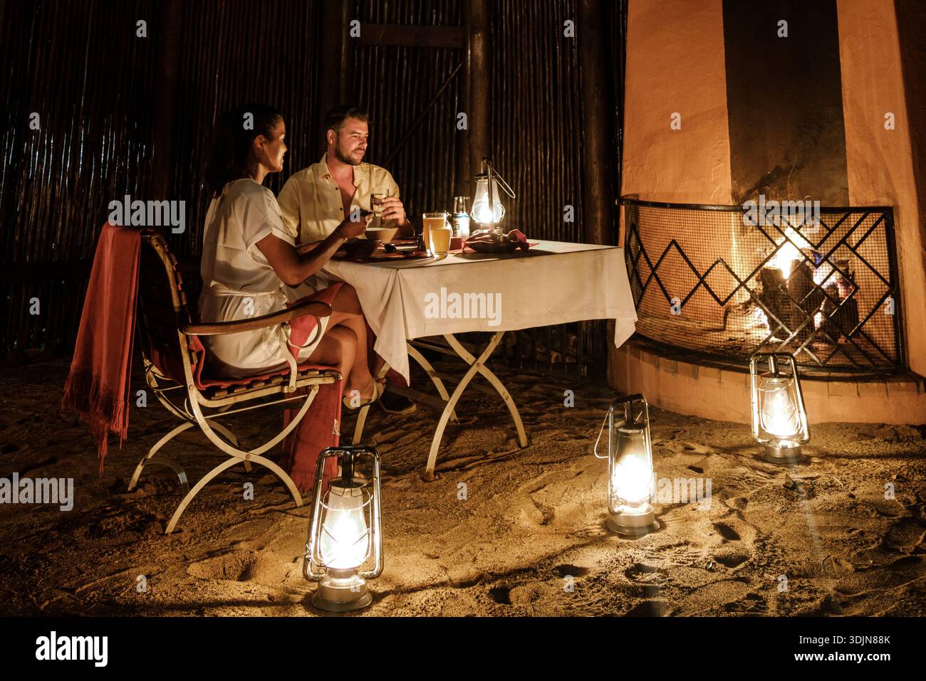 Coppia che gusta una cena intima immersa nella natura nel Parco Nazionale di Kruger, Sud Africa. Il caldo bagliore delle lanterne e un accogliente fuoco esaltano la m Foto Stock