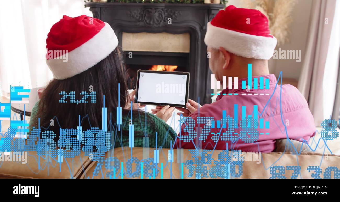 Coppia seduta nei cappelli di Babbo Natale di fronte al caminetto, una tavoletta verde e una rosa che mostra grafici Foto Stock