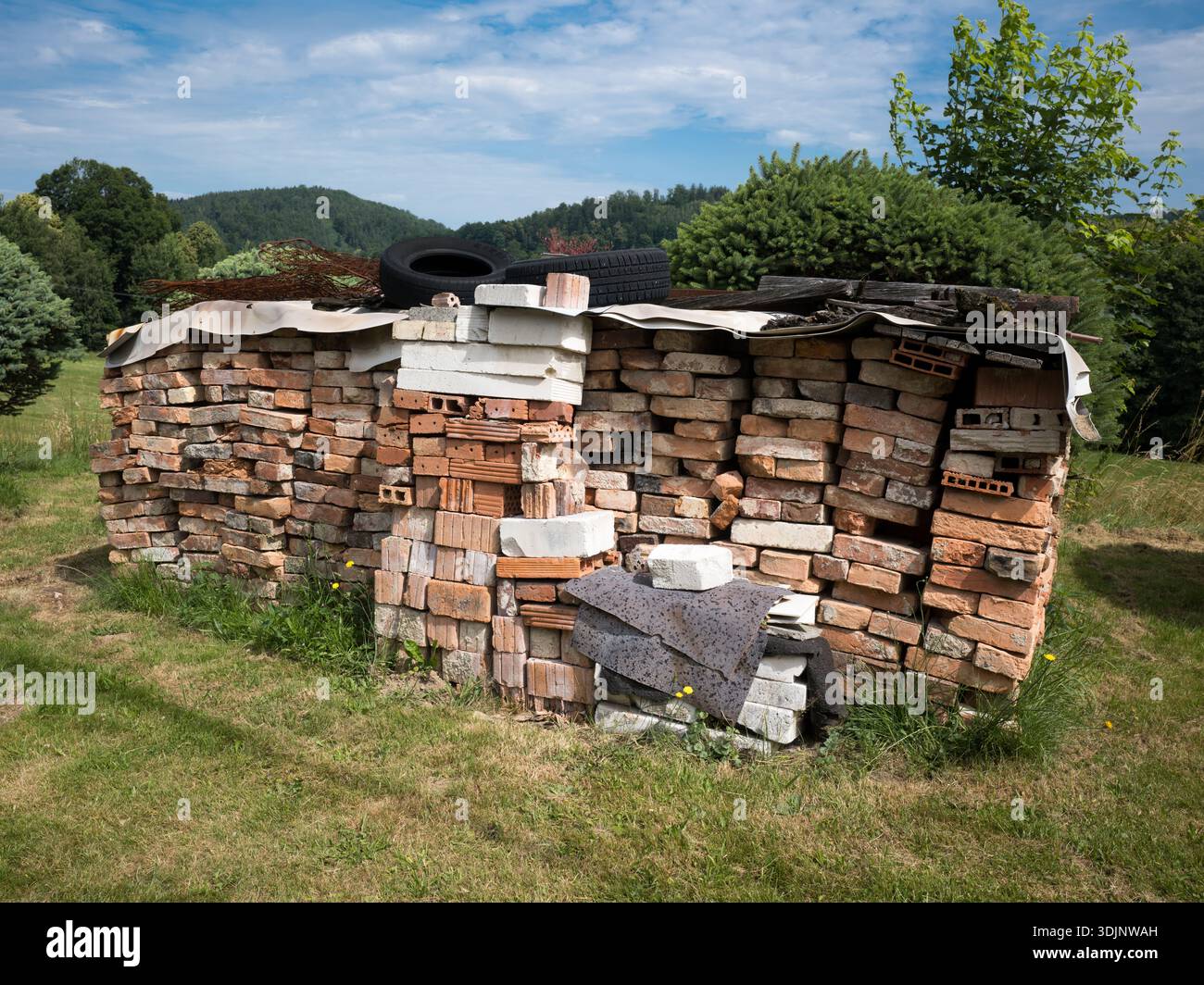 Una grande pila di vecchi mattoni rossi e bianchi ricoperti da un telo e pneumatici, in un campo erboso con verdi colline e alberi sullo sfondo. Foto Stock