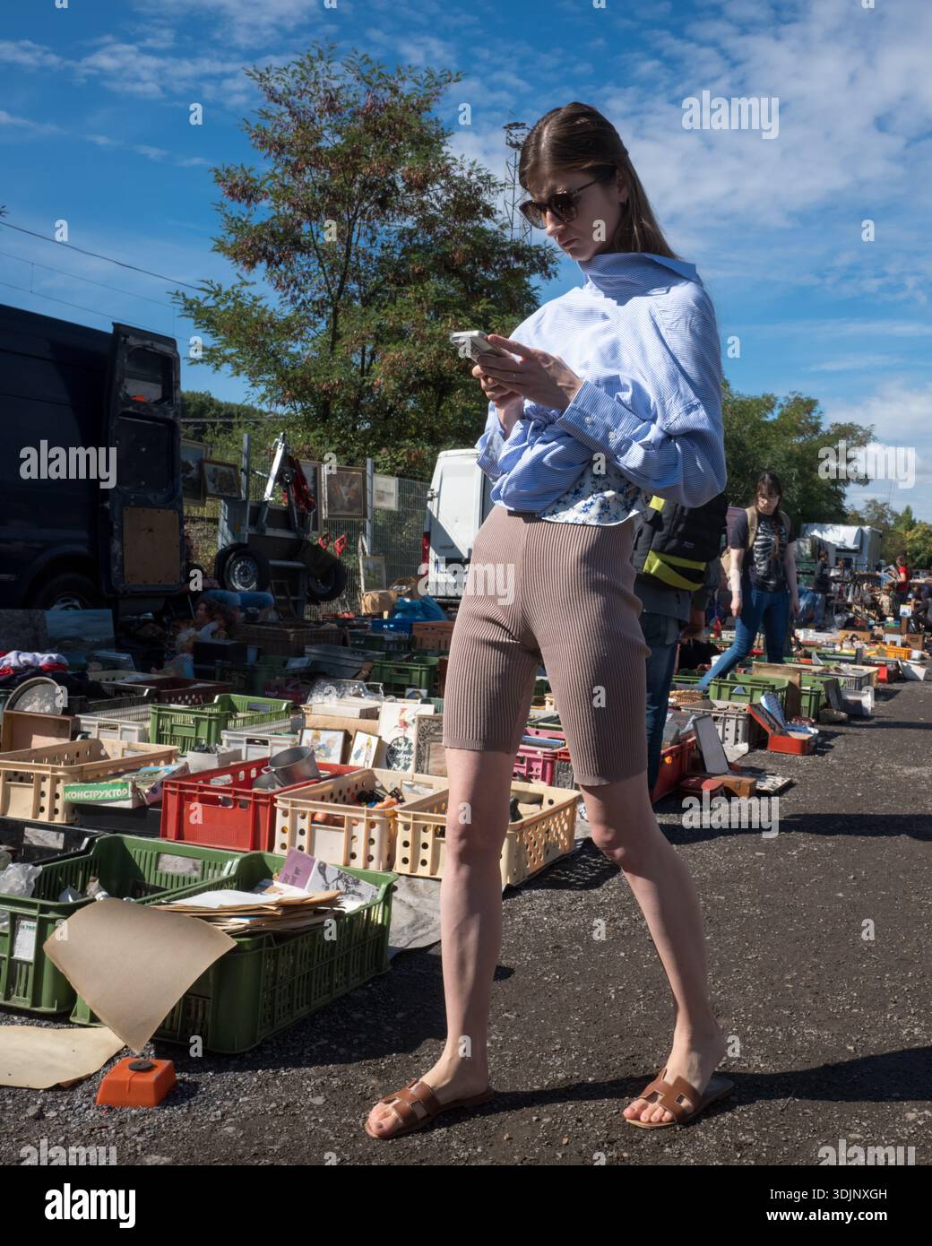 Una giovane donna con occhiali da sole e pantaloncini da motociclista che guarda il suo smartphone mentre cammina attraverso un mercato delle pulci all'aperto con merci in casse di plastica. Foto Stock