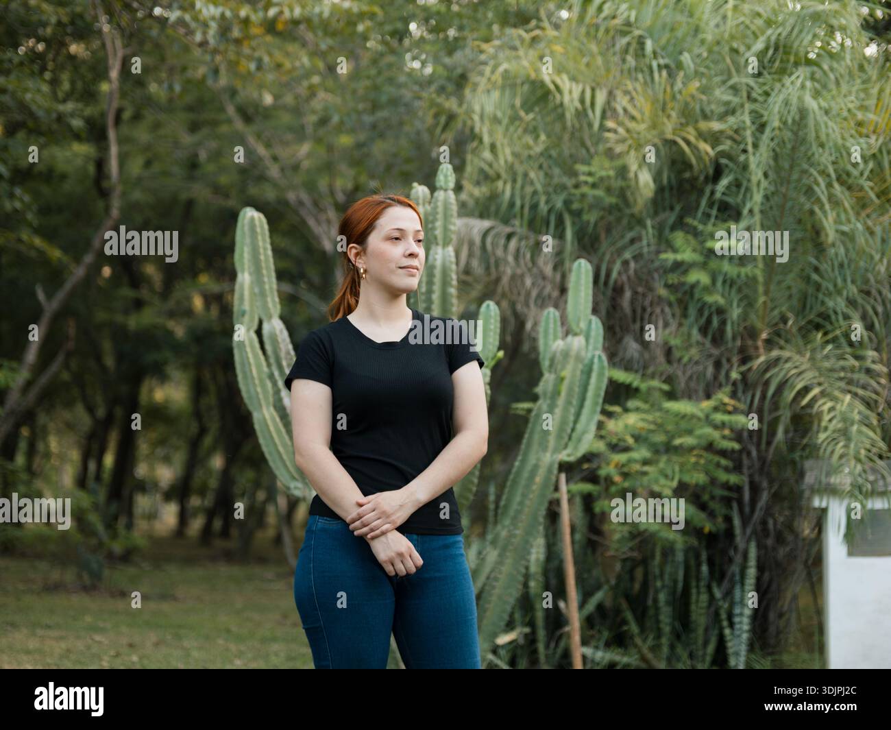 Immagine di una giovane donna in piedi in un parco con cactus e vegetazione sullo sfondo, con una postura rilassata e che guarda di lato Foto Stock