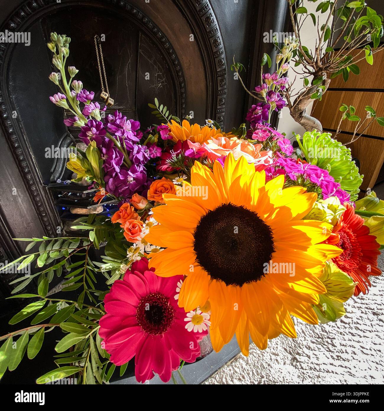Mazzo misto luminoso di girasoli, gerberas, rose e altri fiori colorati disposti di fronte a un tradizionale caminetto in ghisa nera in un ambiente intimo Foto Stock