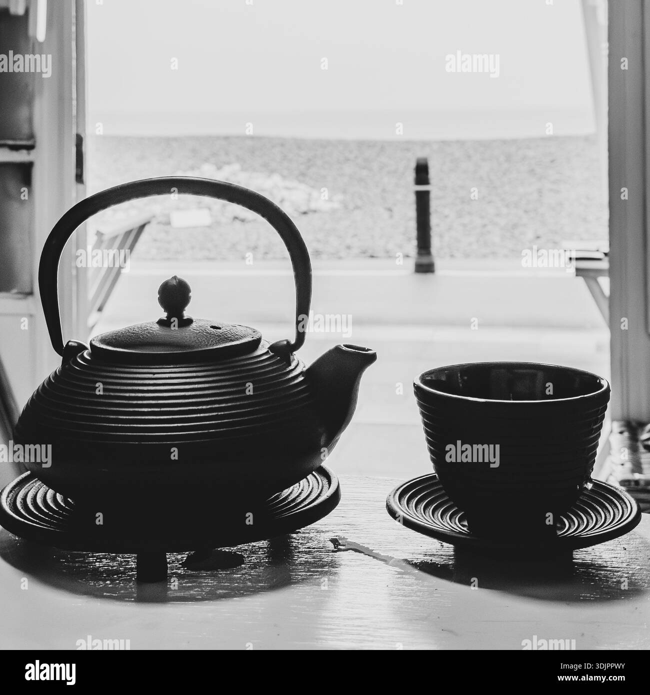 Teiera giapponese in ghisa nera e tazza su un piattino in una cucina sul mare, vista da una porta aperta verso il mare in monocromia. - Immagine stock catturata con smartphone