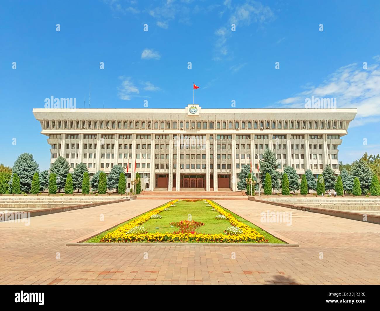 Bishkek, Kirghizistan - 12 giugno 2024: Grande edificio amministrativo con una facciata classica e bandiera, circondato da verde paesaggistico Foto Stock