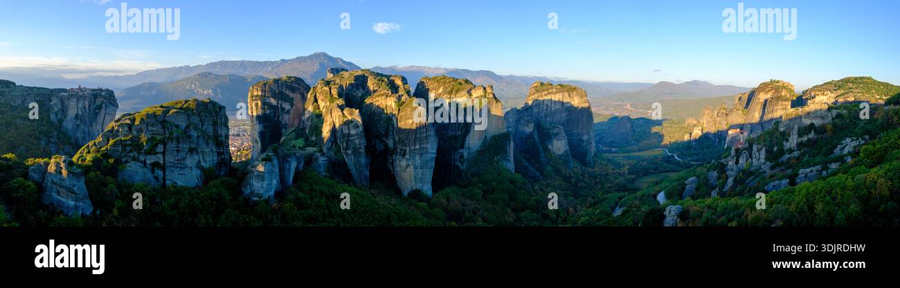 Vista panoramica dei monasteri di Meteora e delle formazioni rocciose che si innalzano sopra una valle boscosa con montagne lontane sotto un cielo blu, Tessaglia, Grecia Foto Stock