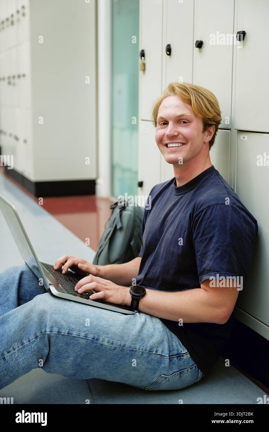 Ritratto di un giovane sorridente seduto con un laptop vicino agli armadietti nel corridoio dell'università Foto Stock