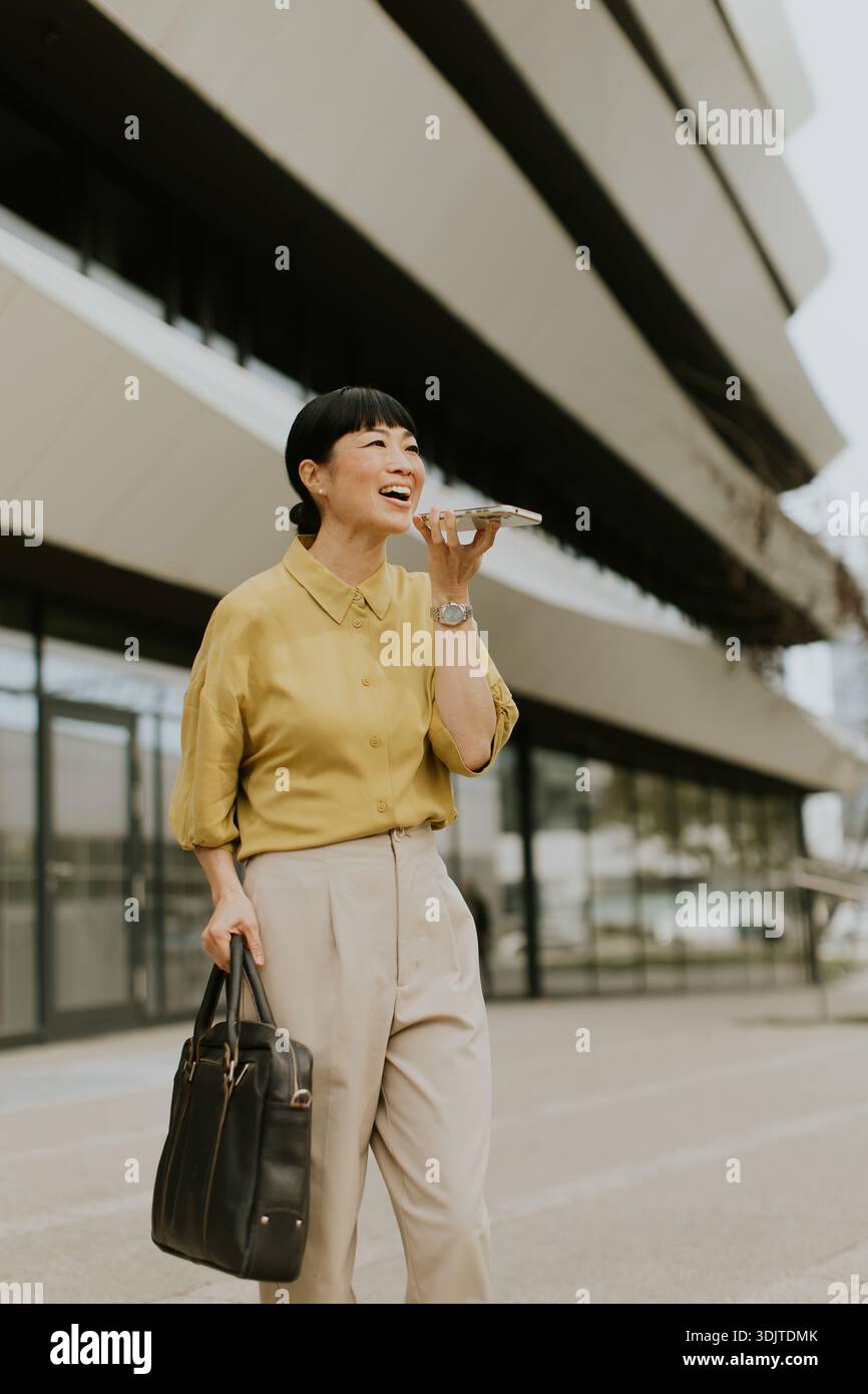 Una donna vestita con una camicetta gialla e pantaloni beige sorride mentre cammina fuori da un moderno edificio per uffici. Tiene uno smartphone in una mano e un bla Foto Stock