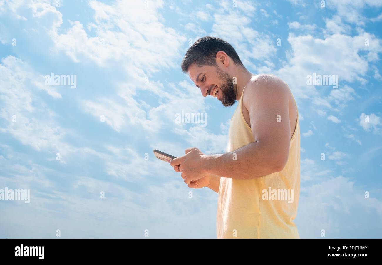 Ridendo lo spagnolo giovane sulla spiaggia scrivendo messaggi con il cellulare e un bel cielo azzurro Foto Stock
