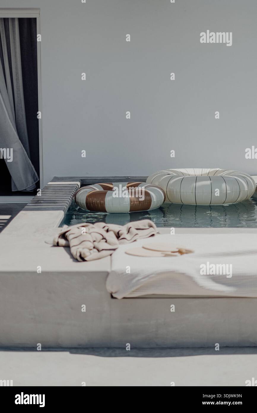 Ambiente minimalista a bordo piscina con anelli gonfiabili, asciugamani bianchi e acqua calma in un moderno ambiente dell'isola greca. «» Foto Stock