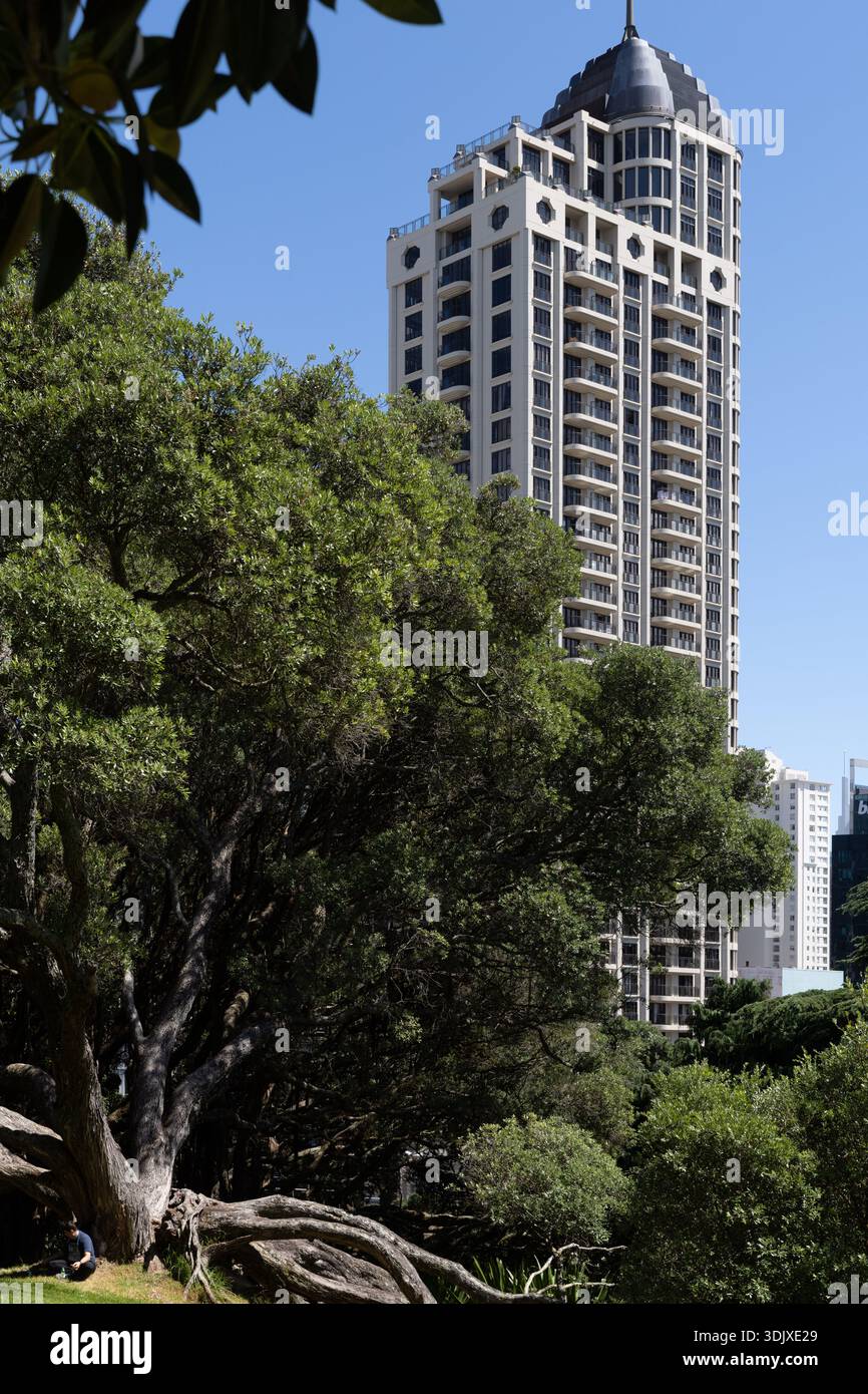Alta torre residenziale che sorge sopra gli alberi nell'Albert Park, Auckland, con una fitta vegetazione che contrasta l'architettura moderna della città. Foto Stock