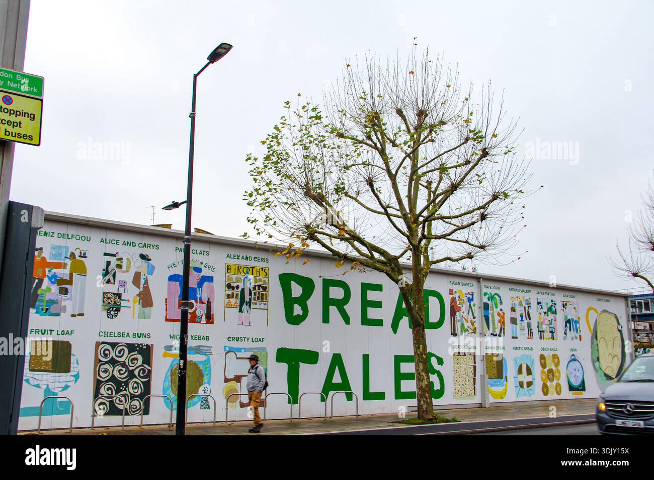 Murale urbano di Street art intitolato "FAVOLE DI PANE" su una parete bianca della città con colorate illustrazioni di panifici, un albero sottile e un passeggio pedonale. Foto Stock