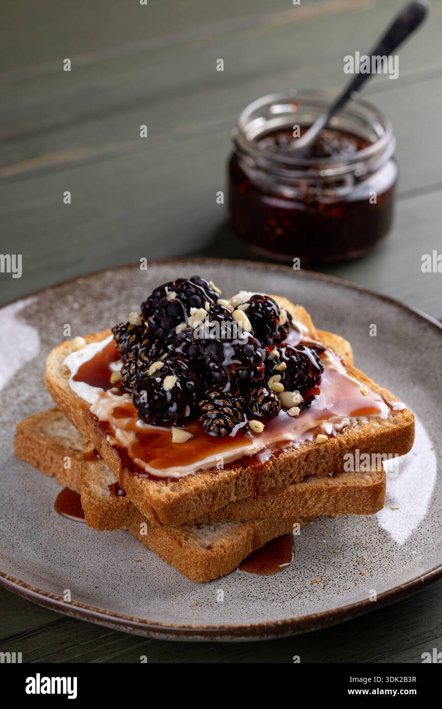 Primo piano di due fette di pane leggero tostato e spalmato con formaggio spalmabile, ricoperto da un mucchio di more fresche, condite con marmellata o sciroppo, a. Foto Stock