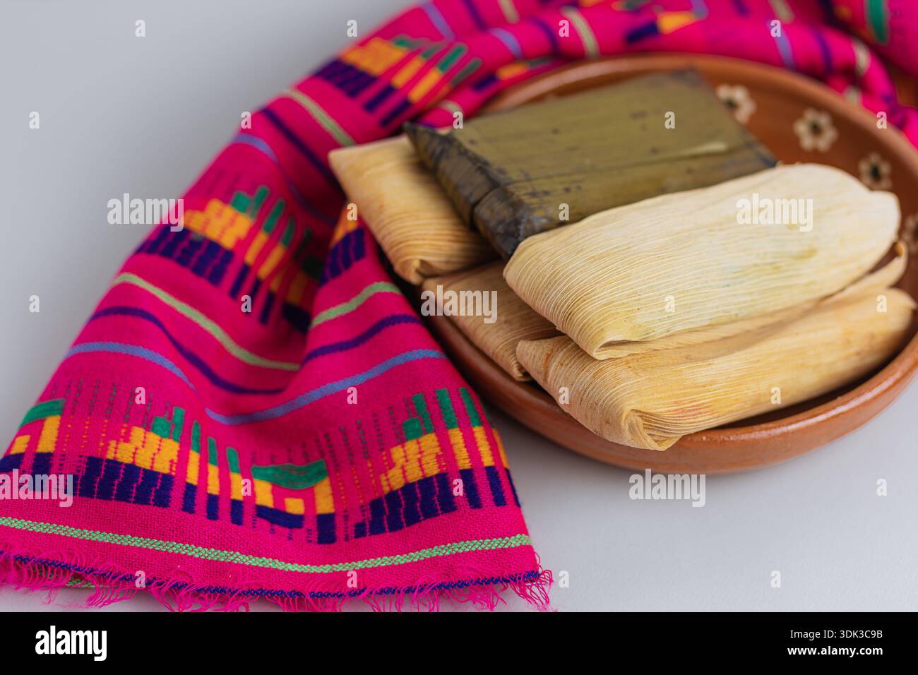 Gruppo di tamales avvolti in bucce di mais e foglie di banana, serviti su un piatto di argilla e colorati tessuti artigianali Foto Stock