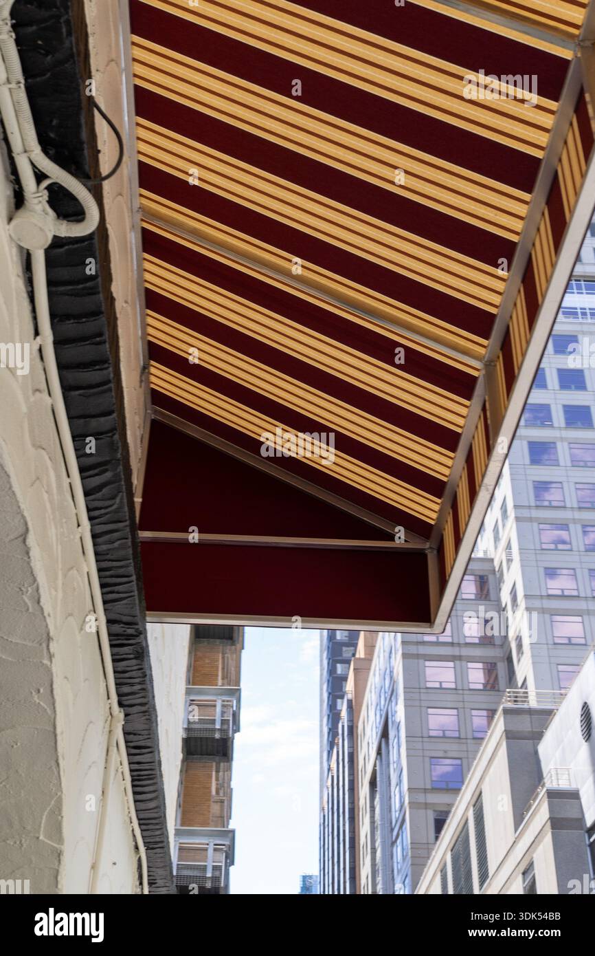 Una vista ad angolo verso l'alto cattura una tenda a strisce che fornisce ombra, incorniciata da un muro testurizzato e torreggianti edifici cittadini sotto un cielo limpido. Foto Stock
