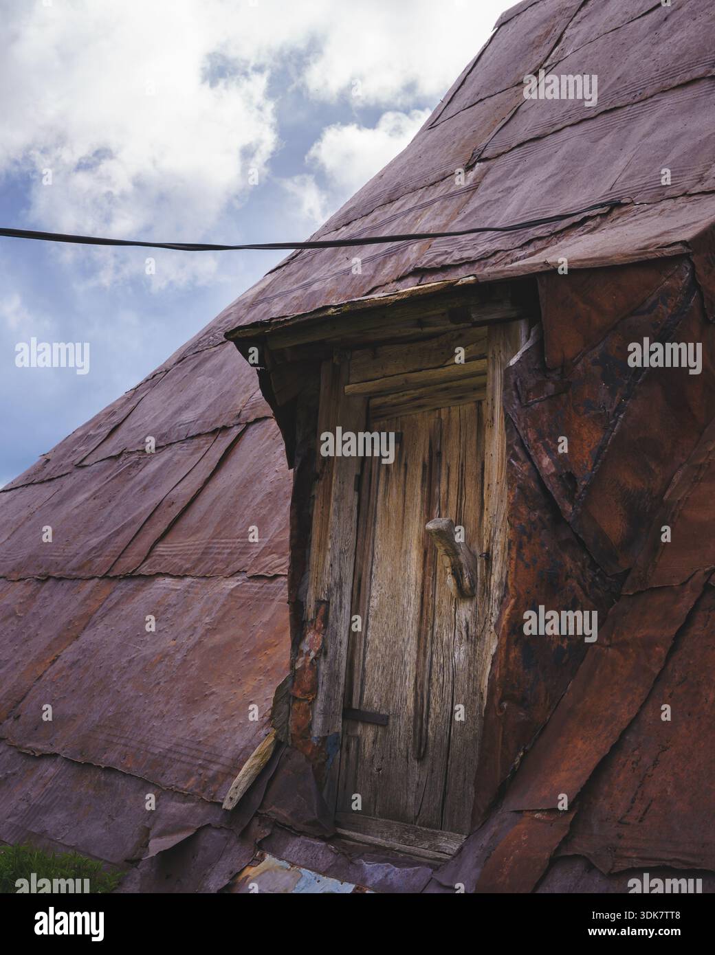 Vista di una porta di legno intemprata all'interno dell'abbraccio arrugginito e testurizzato di un tetto spiovente sullo sfondo di un cielo nuvoloso, Lukomir, Bosn Foto Stock