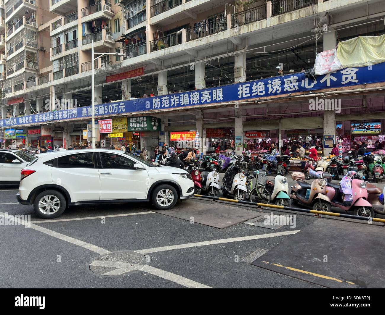 Nanning, Cina - 7 gennaio 2024: Vivace scena di strada in Cina con scooter parcheggiati e negozi che fiancheggiano un edificio a più piani Foto Stock