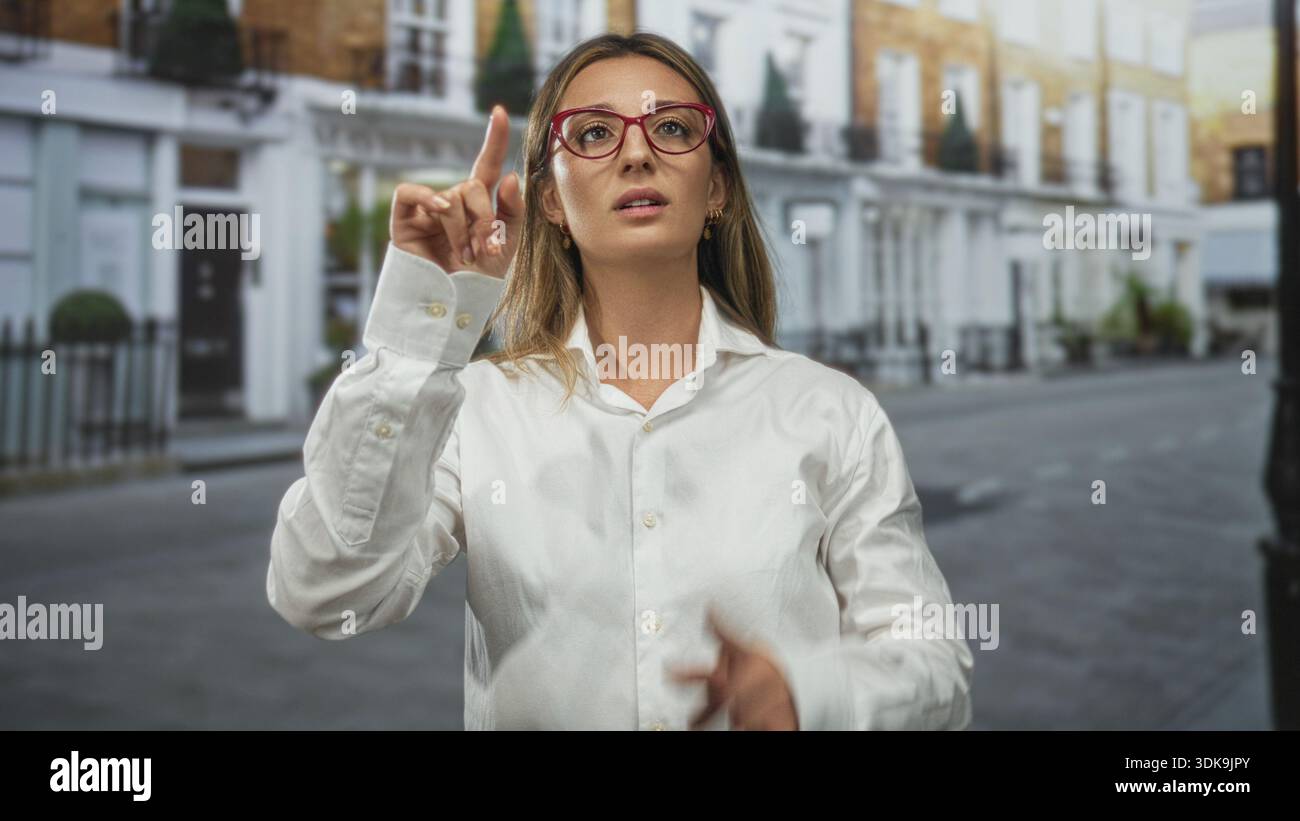 Giovane donna bionda punta il dito verso l'alto sulla strada indossando occhiali rossi e camicia bianca; determinazione. Foto Stock
