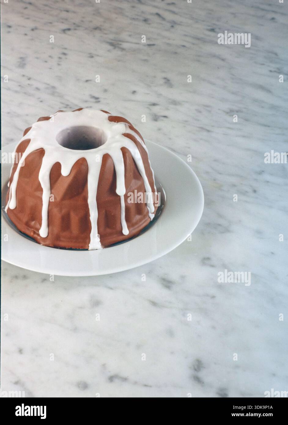 Una torta al cioccolato completa e rotonda ricoperta di liscia glassa bianca, elegantemente presentata su un piatto bianco su uno sfondo di marmo bianco. Foto Stock