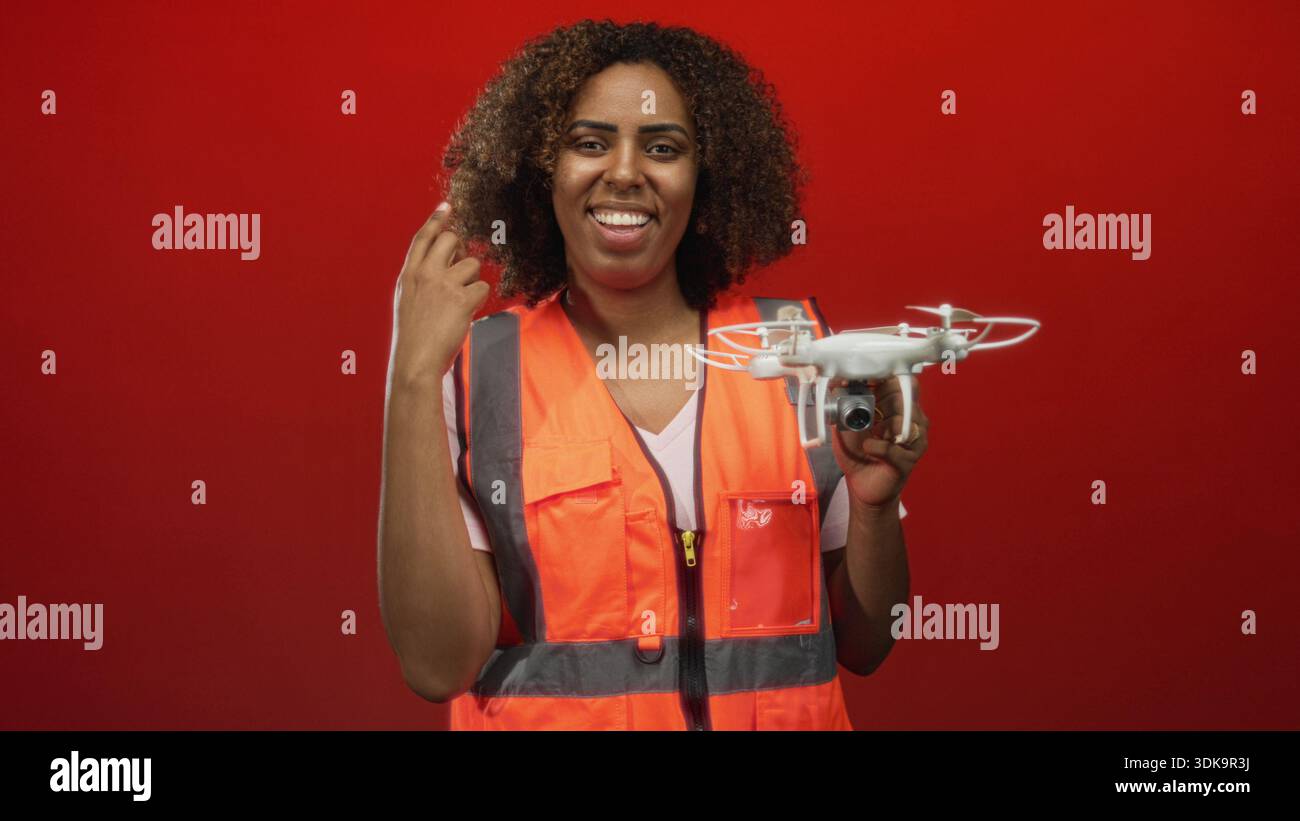 Donna che tiene in mano un drone e fa una pompa a pugno con mano visibile mentre sorride in studio con sfondo rosso; risultato emozionante. Foto Stock